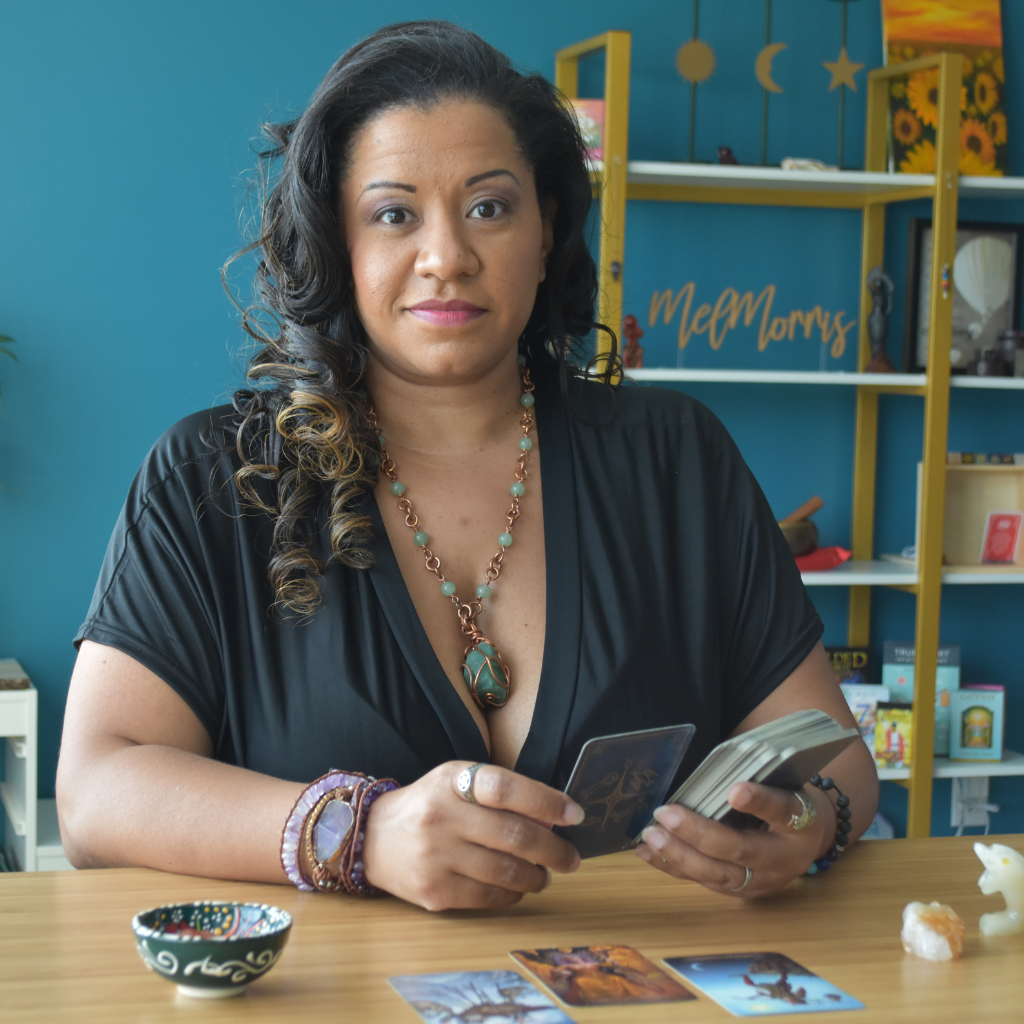 african american woman with long curly hair siting at desk holding tarot cards wearing gemstone jewelry